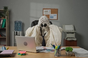 Halloween businessman ghost in white sheet necktie sitting at office desk works with financial papers, tired expression holding head.
