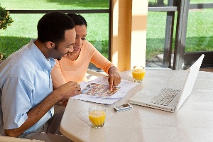 Couple in a blue shirt and orange shirt pointing at flyer and talking. Two glasses of orange juice, cell phone, and laptop open on the table.
