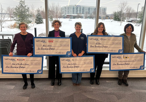Picture of local quad city nonprofits holding big checks from IHMVCU in front of a window.