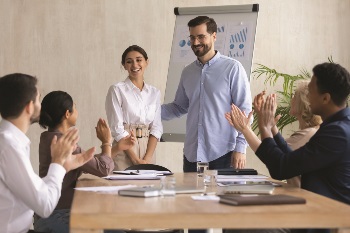 Woman with coworkers, recieving a promotion. Coworkers are clapping and smiling. Man with glasses in blue shirt patting woman on the back in congratulations.