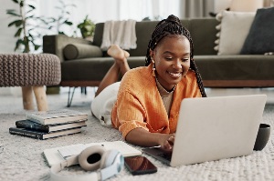 young woman in orange laying on ground with her laptop and electronics surrounding her