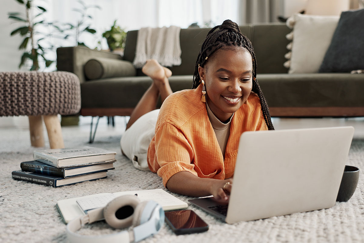 young woman in orange laying on ground with her laptop and electronics surrounding her