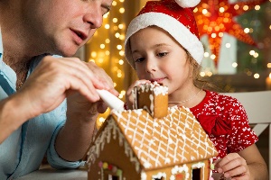 Dad and daughter decorating a gingerbread house with holiday lights behind them