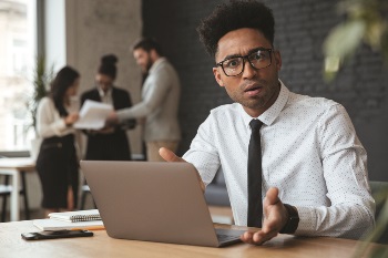 Young black man on laptop at work looking at the camera confused